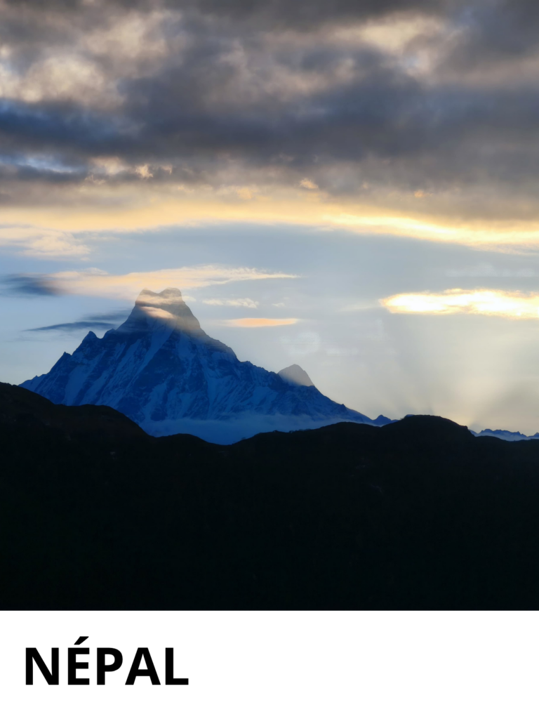 Sommet enneigé de l'Himalaya au Népal émergeant des nuages au lever du soleil, avec des silhouettes de montagnes sombres au premier plan et un ciel dramatique aux teintes dorées et grises